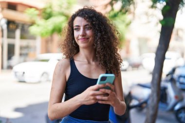 Genç, güzel, İspanyol bir kadın sokakta akıllı telefon kullanarak gülümsüyor.