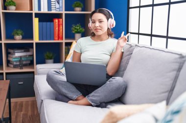 Evde dizüstü bilgisayar kullanan, kulaklık takan, kameraya gülümseyen ve zafer işareti yapan İspanyol bir genç kadın. İki numara.. 