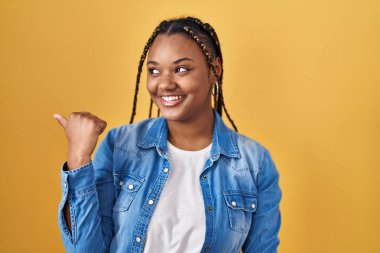 African american woman with braids standing over yellow background smiling with happy face looking and pointing to the side with thumb up. 