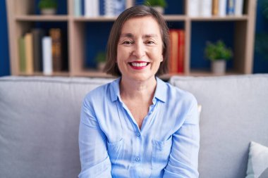 Orta yaşlı İspanyol bir kadın mutlu ve havalı bir gülümsemeyle evdeki kanepede oturuyor. şanslı kişi. 