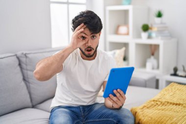 Kanepede oturmuş dokunmatik ped kullanan İspanyol sakallı bir adam şaşırmış yüz ifadesiyle şaşırmış, parmaklarından bakan bir jest yapıyor. İnançsız ifade. 