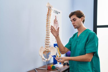 Young caucasian man physiotherapist touching anatomical model of spinal column at rehab clinic