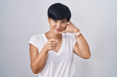 Kısa saçlı genç Asyalı kadın izole edilmiş arka planda duruyor gülümsüyor telefonda konuşuyor ve seni işaret ediyor. Ara beni.. 