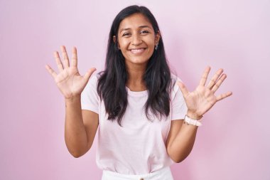 Pembe arka planda duran genç İspanyol kadın on numarayı gösterip işaret ederken kendinden emin ve mutlu bir şekilde gülümsüyor.. 