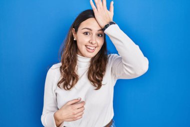 Young hispanic woman standing over blue background surprised with hand on head for mistake, remember error. forgot, bad memory concept.  clipart