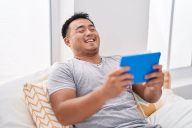 Young chinese man watching video on touchpad sitting on bed at bedroom