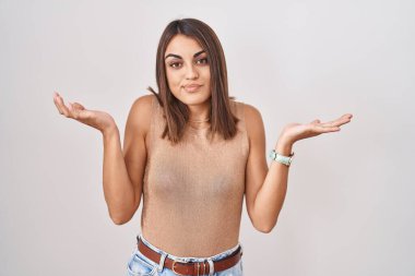 Young hispanic woman standing over white background clueless and confused expression with arms and hands raised. doubt concept. 