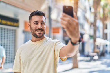 Genç İspanyol adam gülümsüyor. Sokakta akıllı telefonun yanında özçekim yapıyor.