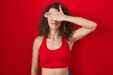 Kıvırcık saçlı İspanyol kadın. Kırmızı arka planda duruyor. Gözlerini eliyle kapatıyor, ciddi ve üzgün görünüyor. Görme, saklama ve reddetme konsepti 