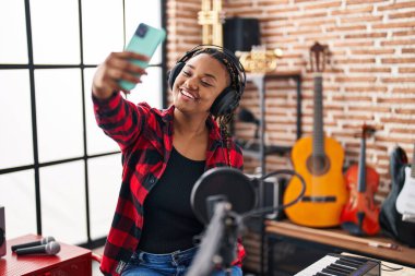 Gülümseyen Afrikalı Amerikalı kadın müzisyen müzik stüdyosunda akıllı telefondan özçekim yapıyor.