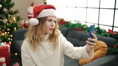 Sarışın bir kadın akıllı telefonuyla selfie çekiyor. Evdeki Noel ağacının yanında koltukta oturuyor.