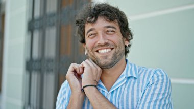 Young hispanic man smiling confident doing heart gesture with hands at street
