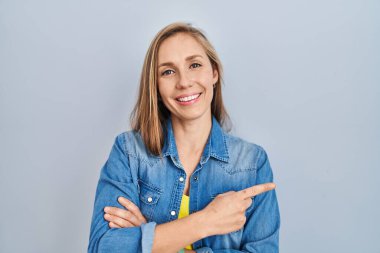 Mavi arka planda duran genç sarışın kadın yüzünde büyük bir gülümsemeyle, kameraya el ile işaret ederek bakıyor.. 