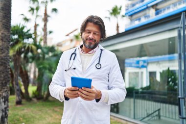 Orta yaşlı doktor hastanede dokunmatik ped kullandığından emin bir şekilde gülümsüyor.