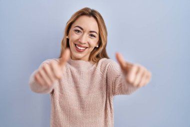Mavi arka planda durup olumlu jestler yapmayı onaylayan İspanyol bir kadın. Gülümsüyor ve başarı için seviniyor. kazanan jest. 