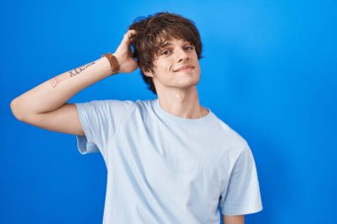 Mavi arka planda duran Latin asıllı genç adam gülümsüyor. Kendine güvenen, el hareketleriyle saçlara dokunan, çekici ve şık poz veren. 