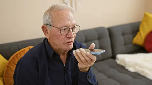 Serious-faced elder man with white hair, engrossed in the technology of a smartphone, sends an audio message from his living room sofa at home