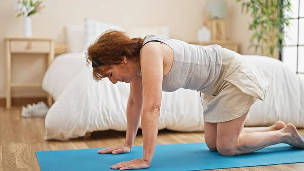 Middle age woman stretching back sitting on yoga mat at bedroom