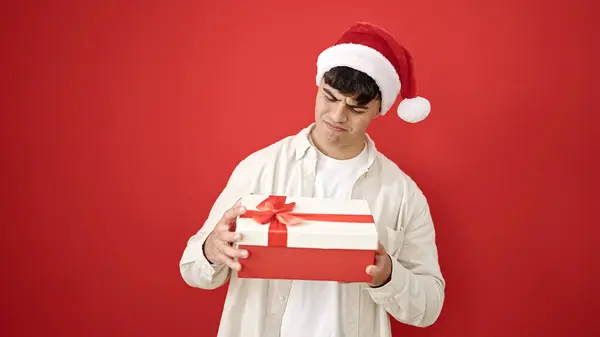 クリスマスの帽子をかぶっている若いヒスパニック男性は 孤立した赤い背景の上に動揺しているように見える贈り物を解放します — ストック写真