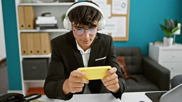 Chillaxed young hispanic work-boss teenager playing video game indoors at the office, tuned in with headphones, absorbed in the gamer\'s world, the background noise of business hustle fading out!