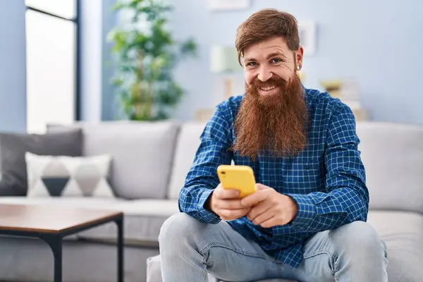 Young redhead man using smartphone sitting on sofa at home