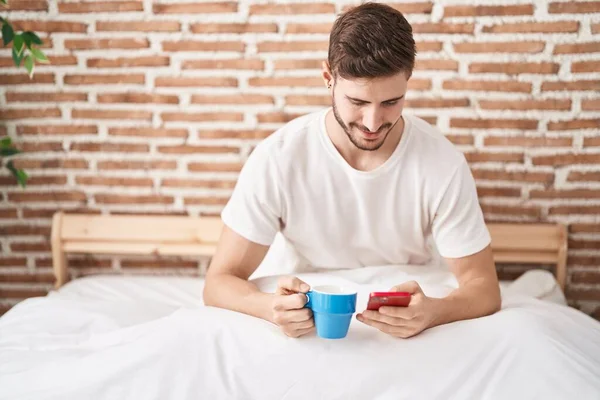 Beyaz bir adam akıllı telefon kullanıyor. Yatak odasında kahve içiyor.