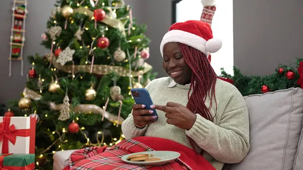 African woman with braided hair using smartphone sitting by christmas tree at home