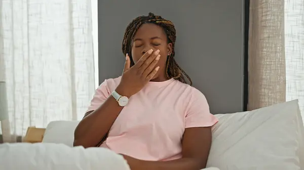 African american woman sitting on bed yawning at bedroom