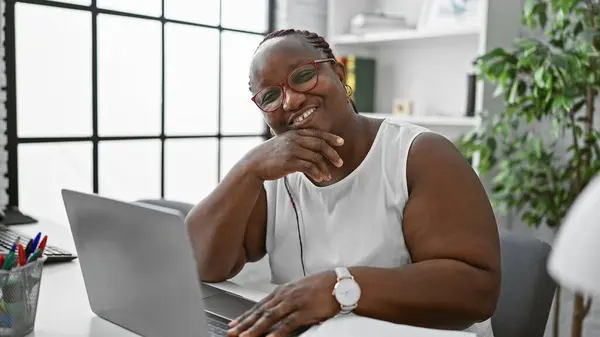自信的非洲裔美国女性老板 摇摇晃晃的辫子和眼镜 微笑着在优雅的办公桌前的笔记本电脑上工作 — 图库照片