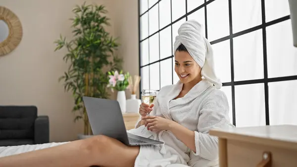 Young beautiful hispanic woman wearing bathrobe drinking champagne having video call at bedroom