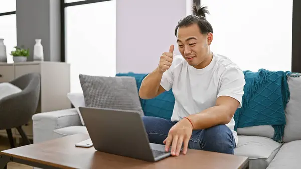 Cheerful young chinese man, pigtails hairstyle, sitting cozily at home, working happily online, using laptop on sofa, flashing a heartening thumb up gesture
