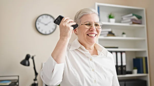 Middle age grey-haired woman business worker listening to voice message by smartphone working at office
