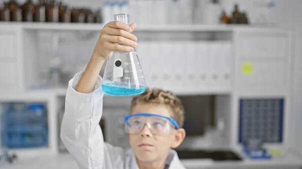 Adorable blond boy scientist totally immersed in experiment, looking at test tube, full of security and focus in lab, little face of science and professional research indoors.
