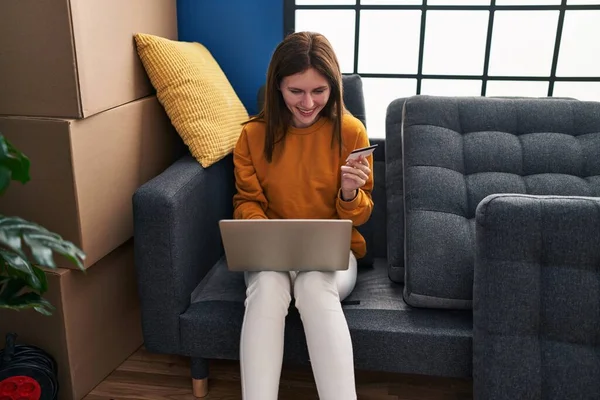 Young blonde woman shopping with laptop and credit card sitting on sofa at new home