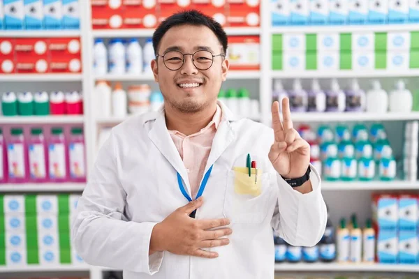 Chinese young man working at pharmacy drugstore smiling swearing with hand on chest and fingers up, making a loyalty promise oath