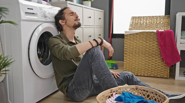 Young hispanic man washing clothes sitting on floor tired at laundry room