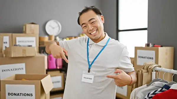 チャリティーセンターでボランティアをしている自信のある若い中国人男性 忙しい屋内倉庫で彼のアイドルバッジを指差しながら笑顔 — ストック写真