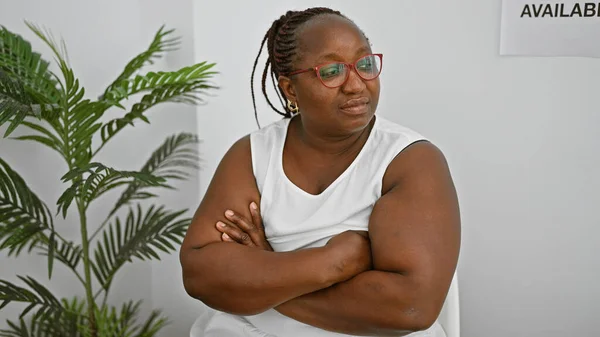 严重的非洲裔美国女人 一位戴着辫子和眼镜的成功的经理 坐在候车室里放松而专注 双臂交叉 细细地审视着周围的环境 — 图库照片