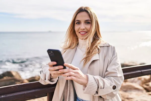 Young blonde woman smiling confident using smartphone at seaside