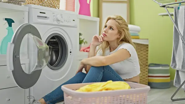 洗濯室で考えている床に座っている若い金髪の女性 — ストック写真