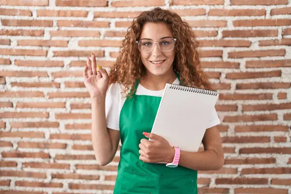 年轻的高加索女人拿着艺术笔记本 微笑积极地做着手指手画脚的手势 成功表达 — 图库照片
