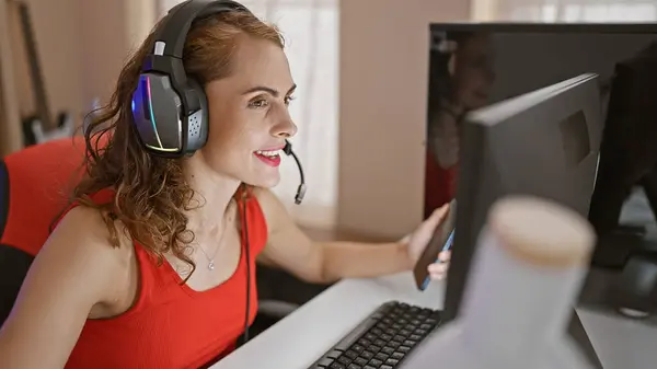 Gaming night unfolds as beautiful caucasian woman streamer syncs smartphone and computer in home office gaming room while smiling, typing, broadcasting her epic plays online