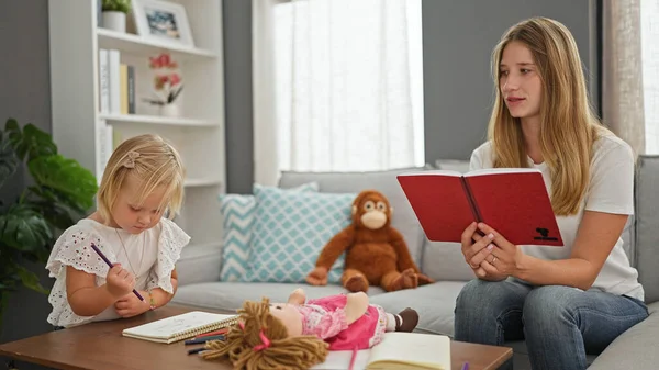 Caucasian mother and her little girl absorbed in a heartwarming storybook at home, joyfully drawing in their notebook amidst a comfortable, familiar indoor setting