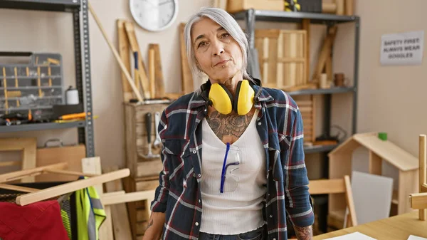 勤劳的中年女性 一个灰白头发 严肃脸蛋的木匠 为自己装备精良的木工车间 耳机的安全和专注而自豪 — 图库照片