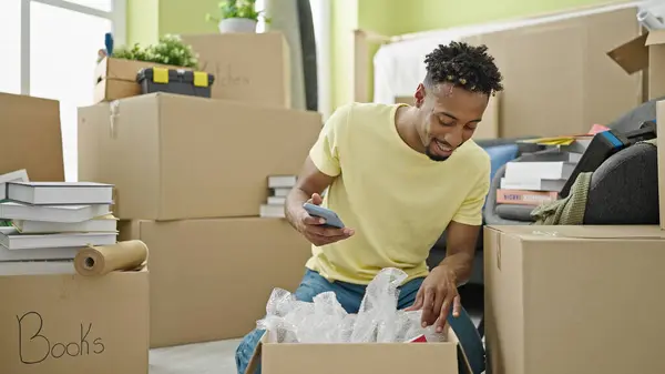 African american man unpacking cardboard box using smartphone at new home