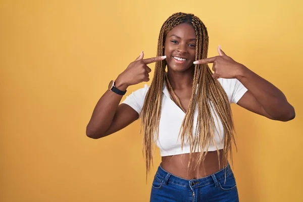 站在黄色背景上 留着辫子的非洲裔美国女人开朗地微笑着 用手指和嘴指点着 牙齿健康概念 — 图库照片