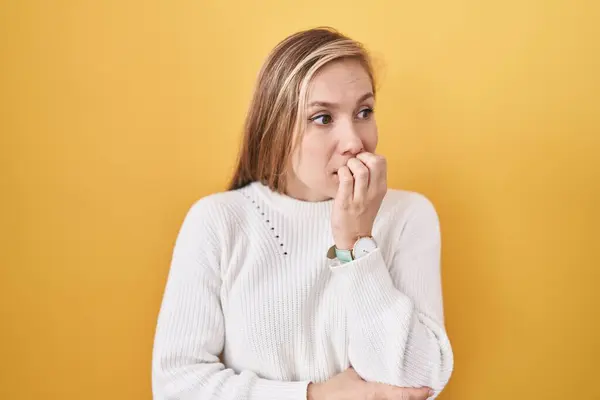 黄色の背景に白いセーターを着用した若い白人女性は ストレスを感じて 爪を噛む手で緊張しています 不安問題 — ストック写真