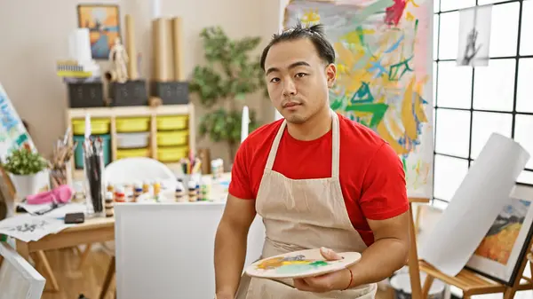 Serious-faced young chinese man, an emerging artist engrossed in his painting, sits on a studio chair, canvas and paintbrush in hand, absorbed in the world of art and creativity.