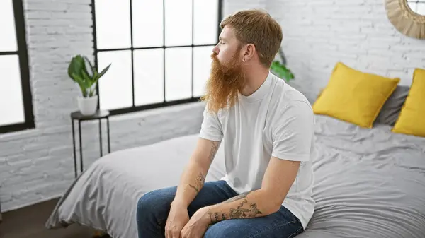 Handsome, young redhead man comfortably sitting on his bed in a relaxed morning, intentionally looking to the side with a serious expression on his face in his cozy bedroom.