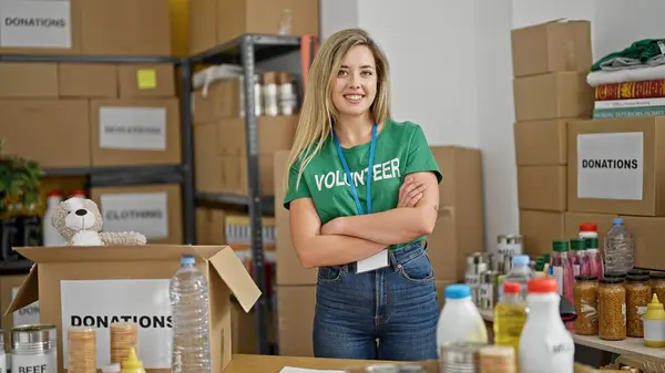 Young blonde woman volunteer standing with arms crossed gesture smiling at charity center